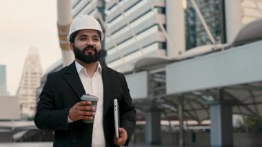 Kasklı mutlu Hintli mühendisin portresi. Elinde kahve fincanı ve açık ofis binaları var. Ofis kostümü konusunda uzman bir endüstriyel uzman cadde boyunca yürüyor. Sanayi işi