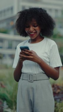 Megapolis caddesinde akıllı telefondan sohbet okuyan mutlu Afro-Amerikan kadın portresi. Şehir ortamında cep telefonu ekranında neşeli kadın mesajları. Yaşam tarzı dikey çekim