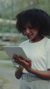 Portrait of pretty African American lady browsing internet on tablet computer outdoors. Interested female checks social media on digital device in city park. Vertical shot