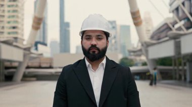 Çağdaş binaların önünde duran kasklı Hintli erkek müteahhit portresi. Ceketli, sakallı endüstriyel müfettiş sokaklara güvendiğini ifade ediyor.