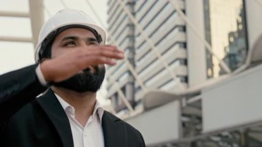 Yükselen binalara bakan güneşten gözlerini kapayan miğferli Hintli yöneticinin portresi. Sakallı müteahhit dışarıda modern gökdelenlere karşı duruyor.