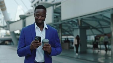Genç Afrikalı Amerikalı menajerin portresi sokakta elinde kahve fincanı tutan akıllı telefona bakıyor. Şirket çalışanı, şehir merkezindeki tatilde meslektaşının mesajlarını okuyor.