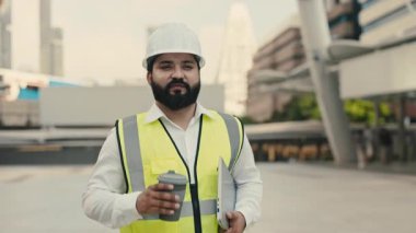 Şehir meydanında yürürken dizüstü bilgisayarı ve kahveyi tutan Hintli mühendisin portresi. İçki ve bilgisayar tebessümleri olan dost canlısı teknisyen inşaat alanında vardiyaya hazır.