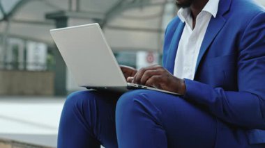 Afrikalı Amerikalı girişimcinin yakından görüntüsü şehir parkında oturan dizüstü bilgisayarla ilgili makale yazıyor. İşadamı şehir ortamında yakın planda belge üzerinde çalışıyor. Uzak görev