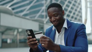 Mutlu Afro-Amerikan iş adamının portresi, şehir merkezindeki akıllı telefonlarda bankacılık uygulaması kullanıyor. Genç girişimci internet teknolojisiyle ödeme yapıyor. Gökdelen yakınındaki korkulukta oturuyor.