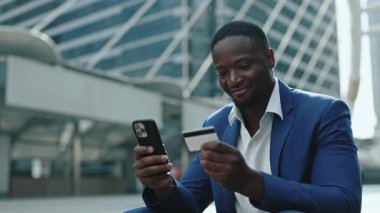 Gülümseyen Afrikalı Amerikalı girişimci portresi şehir bölgesinde akıllı telefon ve kredi kartı bulundurur. Genç yönetici, ofis binasının yanında oturarak güvenli online para transferini seçti