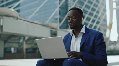 Pozitif Afro-Amerikalı iş adamının portresi laptopla caddede ortakları selamlıyor. Şirket çalışanı gökdelen binasına karşı oturarak video toplantısına katıldı