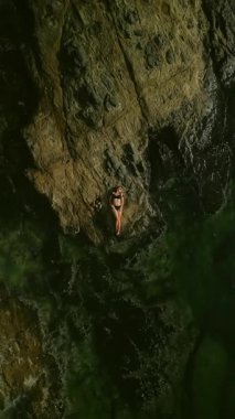 Bikinili kadın turist, büyülenmiş berrak su üzerinde dik kayalık sahilde dinleniyor. Dişi, sakin deniz lagününün yanındaki eski uçurumda yatıyor. Phuket Resort 'un harika doğası. Dikey video