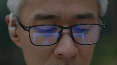 İşitme cihazı açık havada dizüstü bilgisayarda çalışan olgun Asyalı iş adamının yakın plan portresi. Engelli Koreli şirket çalışanı gözlüğe yansıyan dizüstü bilgisayar kullanıyor