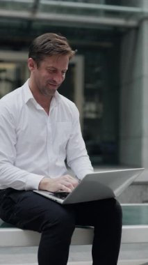 Portrait of smiling man manager works on laptop as sits near corporate building. Mature businessman writes report about successful sales on notebook laptop on street vertical shot