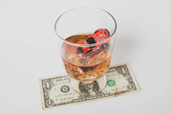A red car is drowning in a glass of alcohol on a dollar bill. mac-up. Social advertising. Concept of drunk driving, car accident, car finance expenses