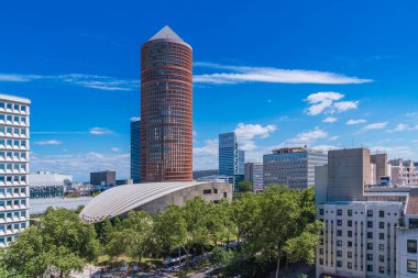 Çarpıcı bir gökdelen, yemyeşil ve diğer binalarla çevrili çağdaş mimariyi gün boyunca açık mavi gökyüzüne karşı sergiliyor..