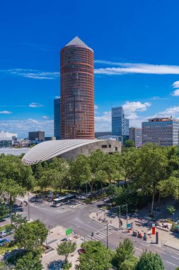 Çarpıcı bir gökdelen, yemyeşil ve diğer binalarla çevrili çağdaş mimariyi gün boyunca açık mavi gökyüzüne karşı sergiliyor.