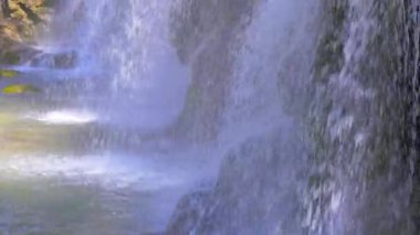 Bir nehir kaynağı kayaların üzerinde çağlayarak güneş ışığında parlayan bir şelale oluşturur.