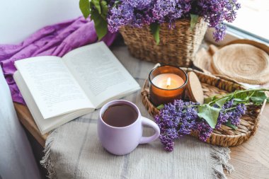 Bir fincan çay, hasır şapka, açık kitap ve leylak sepeti, bahar natürmort hayat.