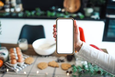 Elinde boş ekranlı telefon, arka planda Noel ışıkları, şenlikli zencefilli kurabiyeler, anason, tarçın, kozalak, kırsal masada sedir dalları. Mesaj için yer var. Mutlu Noeller.