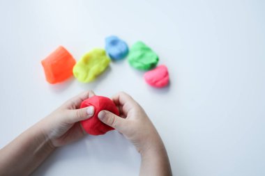 Bebek elleri beyaz masa arkasında kırmızı modelleme kilini tutuyor ve yoğuruyor. - Yakın çekim. Bebek gelişimi. Farklı rengarenk şekiller yapmak için malzeme hazırlanıyor.