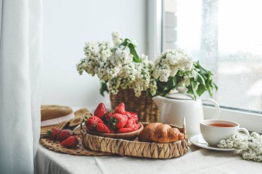 Bir fincan çay, kruvasan, çilek ve leylak sepeti, yaz sabahı estetiği..