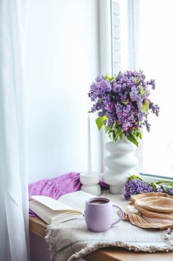 Bir fincan çay, hasır şapka, açık kitap ve leylak sepeti, bahar natürmort hayat.