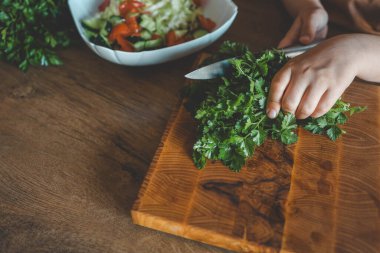 Bir çocuk sebze doğruyor, yapraklı sebzeler ve otlar gibi, ahşap bir kesim tahtasının üzerinde, mutfaktaki lezzetli bir tarifin malzemelerini hazırlamak için.