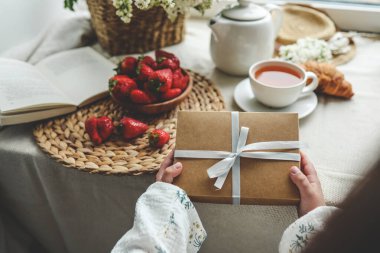 Çocuğun elleri güzel bir hediye kutusunu tutuyor. Arkaplanda bir kurdele ve şenlikli bir kahvaltı var. Üst manzara, yakın plan. Anneler günün kutlu olsun..