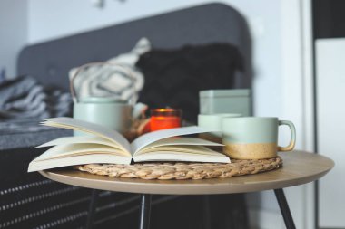 Açık bir kitap, bir fincan çay, bir çaydanlık ve yatak odasının arka planında bir mum..