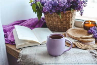 Bir fincan çay, hasır şapka, açık kitap ve leylak sepeti, bahar natürmort hayat.