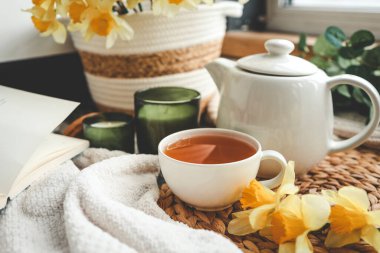 Bir fincan çay, mumlar, açık kitap nergisli sepet, bahar estetik fotoğrafı..