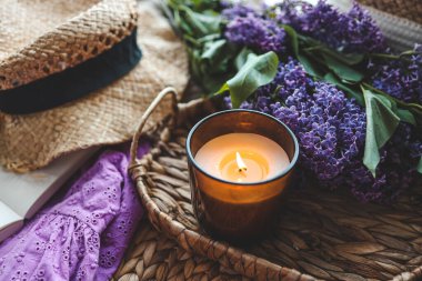 Yanan mum, açık kitap, leylak demeti ve şapka, bahar estetik fotoğrafı..