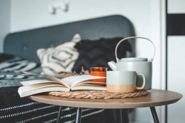 Açık bir kitap, bir fincan çay, bir çaydanlık ve yatak odasının arka planında bir mum..