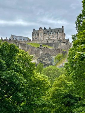 Şehir parkındaki yeşil ağaçların arasından Edinburgh Kalesi 'ne bakıyorum. Arkaplanda kasvetli bulutlar olan yemyeşil bir arkaplan. 