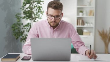 Gözlüklü beyaz bir erkek öğrenci. Masada oturup öğretmenini dinlerken uzaktan video kaydediyor ve deftere bir şeyler yazıyor.