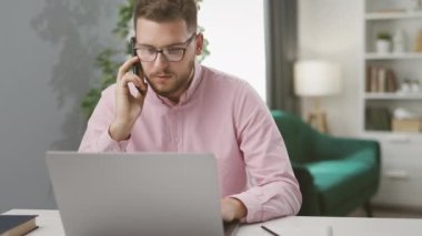 Genç beyaz iş adamı laptopta çalışıyor ve iş ortağıyla akıllı telefondan konuşuyor, uzaktan kumandalı.