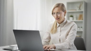 Konsantre sarışın, orta yaşlı, serbest çalışan bir kadın dizüstü bilgisayar kullanıyor, uzaktan kumandalı.