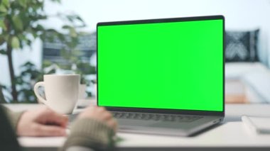 Evden dizüstü bilgisayarla çalışan bir kadın, kroma kopyalama alanı olan bir bilgisayar ekranı, bir fincan dumanı tüten kahveyle samimi bir iç mekan.