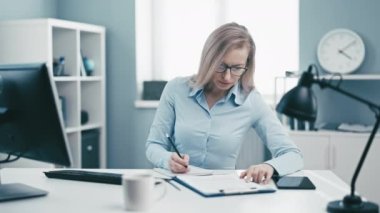 Ofiste oturan olgun bir kadın, masabaşı işyerinde çalışan sarışın bir iş kadını bilgisayardaki belgeleri kontrol ediyor.