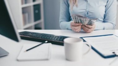 Resmi kıyafetli kadın, ofis masasında çalışan aletlerle dolar sayan banknotlar sayıyor.