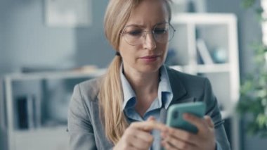 Ofiste akıllı telefon kullanan zeki bir iş kadını. Gözlüklü sarışın bir bayanın finansal bilgilerini kontrol ediyor.