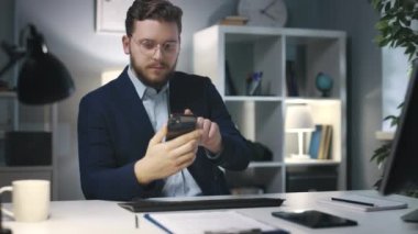 Genç ve başarılı bir iş adamının resmi kıyafet ticareti yapan akıllı telefon ve sakallı bir adamla ofis aletleri kullanarak bilgi kontrol etmesi.