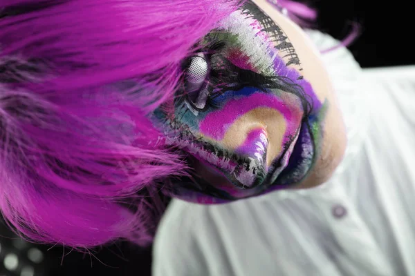 Close up view of woman with purple hair and evil clown face art on black background