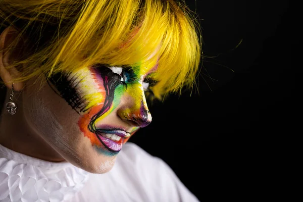 Close up sideway view of woman with yellow hair and evil clown face art on black background