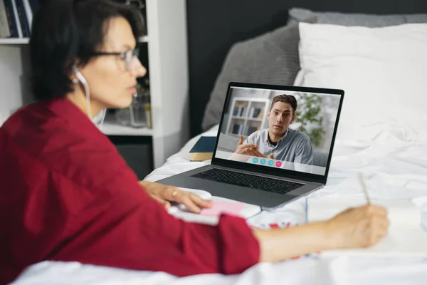 ノートパソコンでのビデオ会議中にメモを取る眼鏡やイヤホンで成熟した女性の側面図 コーカサスのブルネットはベッドルームに座って ビジネスチャットのためにコンピュータを使用して — ストック写真