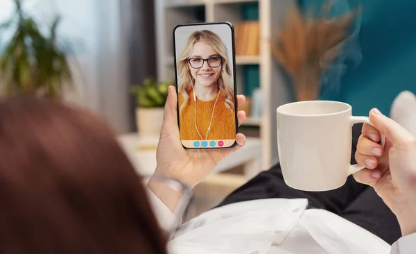 Mature woman lying on comfy couch holding cup of coffee and smartphone with video call, pleasant conversation with daughter. Distancing family video chat