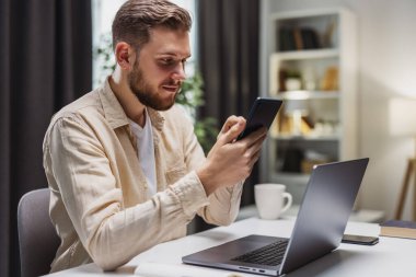 İşadamı akıllı telefonu kaydırıyor, dizüstü bilgisayarla masada oturuyor. Erkek schecking sosyal ağları.