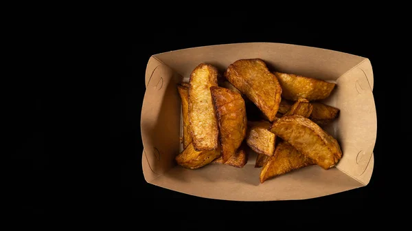 Top view of baked potato slices in paper box, rustic style fast food on black background