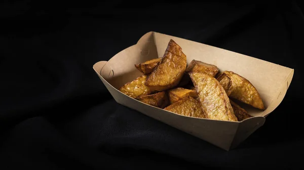 Baked potato slices in paper box, rustic style fast food on black background