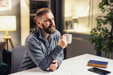 Sakallı, olgun bir adam oturma odasındaki masada oturup bir fincan sıcak kahve ya da çayla rahatlıyor.
