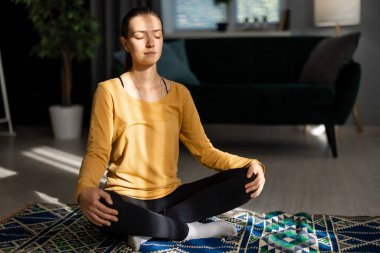 Olgun bir kadın yerde dinleniyor, Lotus 'ta oturuyor. Evde güneş ışınlarına, dinlenmeye ve yoga konseptine karşı poz veriyor.