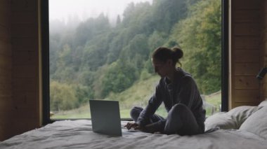 Yetişkin bir kadın dizüstü bilgisayar kullanarak yatakta oturuyor, sisli bir sabah pencerenin arkasında, sakin bir kır evinden çalışıyor.