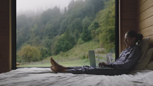 カントリーハウスで働く大人の女性 広い窓付きのベッドルームからのラップトップを使用して 森の霧の朝 落ち着いたレジャーの時間の概念 — ストック動画